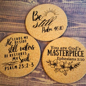 May include: Three round cork coasters with black script. One says "Be still" with a sun graphic and "Psalm 46:10." Another says "He restores my soul Psalm 23:2-3." The third says "You are God's MASTERPIECE Ephesians 2:10."