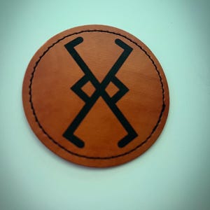 May include: A round, brown leather patch with a black geometric design in the center. The patch has a stitched black border. The design is a series of connected lines and diamond shapes.