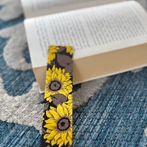 May include: A rectangular bookmark featuring a sunflower design with yellow petals and brown centers against a black background. The bookmark is placed in an open book with white pages, resting on a blue patterned surface.