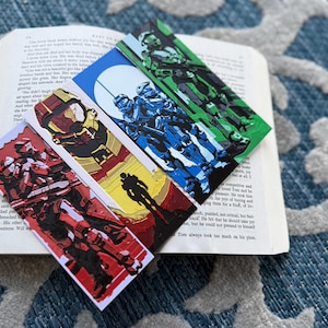 Puede incluir: Cuatro marcadores rectangulares coloridos con ilustraciones de personajes de un videojuego de ciencia ficción, colocados sobre un libro abierto. Los marcadores muestran imágenes en rojo, amarillo, azul y verde.