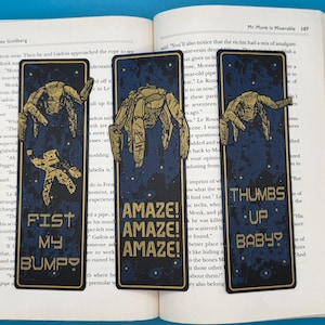 Könnte beinhalten: Drei marineblaue Lesezeichen mit goldenen Illustrationen einer Kreatur und Text. Die Lesezeichen lauten "FIST MY BUMPY", "AMAZE! AMAZE! AMAZE!" und "THUMBS UP BABY?"