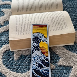 May include: A rectangular bookmark featuring a stylized depiction of a large ocean wave with a setting sun. The wave is rendered in shades of blue and white, with a yellow and red sun. The bookmark is placed on an open book.