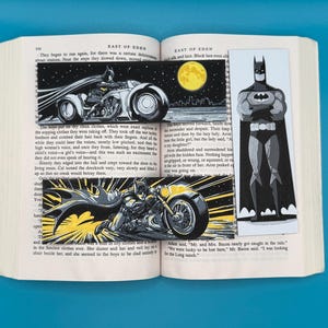 May include: Open book with Batman-themed bookmarks. One bookmark features Batman on a motorcycle in a comic book style. Another shows Batman standing with a cape. The book is open to a page with text.