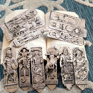 May include: A collection of detailed, gray-toned zodiac sign bookmarks, featuring Aries, Scorpio, Capricorn, Sagittarius, Libra, and Pisces. The bookmarks are placed on an open book, with additional zodiac signs visible.