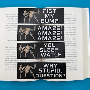 May include: A stack of four black rectangular bookmarks with white text and a beige robot illustration. The text on the bookmarks reads: "FIST MY BUMP", "AMAZE! AMAZE! AMAZE!", "YOU SLEEP. I WATCH.", and "WHY STUPID, QUESTION?"