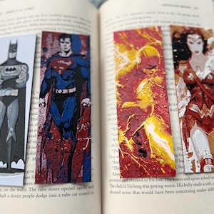 Może przedstawiać: Cztery zakładki do książek z motywem superbohaterów przedstawiające Batmana, Supermana, Flasha i Wonder Woman. Zakładki są prostokątne z szczegółowymi ilustracjami postaci. Zakładki są umieszczone w otwartej książce.