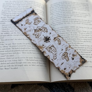 Może przedstawiać: Ręcznie robiona zakładka do książki z motywem mapy i różą wiatrów. Zakładka ma prostokątny kształt z teksturowaną, przypominającą korę krawędzią. Mapa przedstawia brązowe lądy na białym tle. Zakładka leży na otwartej książce.