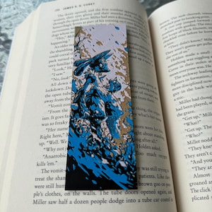 Könnte beinhalten: Ein Lesezeichen mit einer blau-weißen Illustration einer Kreatur. Das Lesezeichen ist rechteckig mit schwarzem Rand und befindet sich in einem offenen Buch mit gedrucktem Text.