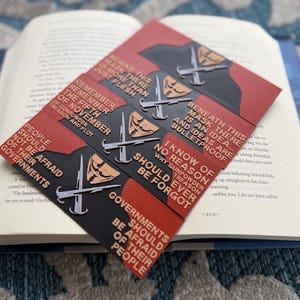 Pode incluir: Um conjunto de marcadores de livros vermelhos e pretos com citações e imagens do filme "V de Vingança". Cada marcador apresenta um design estilizado de máscara e adaga, com texto branco. Os marcadores estão em um livro aberto.