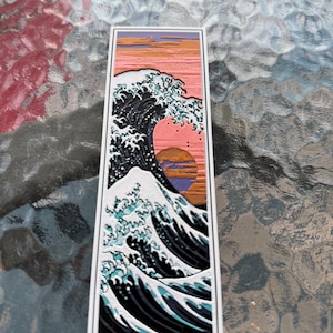 May include: Rectangular sticker featuring a stylized depiction of a large ocean wave, with a sunset in the background. The wave is rendered in shades of black, white, and teal, set against a pink and orange sky. The sticker has a white border.