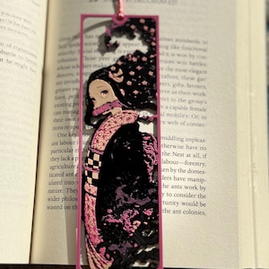 May include: A pink rectangular bookmark featuring a character illustration from an anime series. The character has long black hair and is wearing a pink kimono. The bookmark is inserted into an open book.