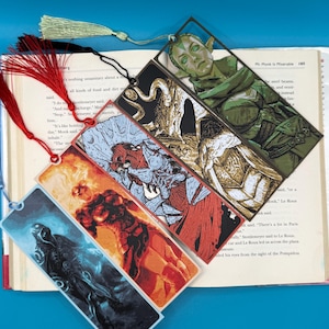 May include: A collection of fantasy-themed bookmarks with tassel accents. Each bookmark features a different character illustration in vibrant colors, including a green-skinned figure, a fiery character, and a shadowy figure.