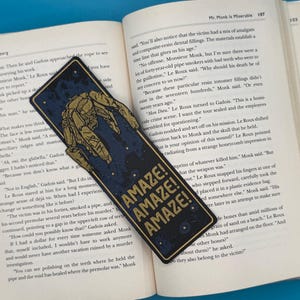 Rocky alien bookmark in dark blue with a gold mechanical outline and AMAZE AMAZE AMAZE text on an open sci fi book.