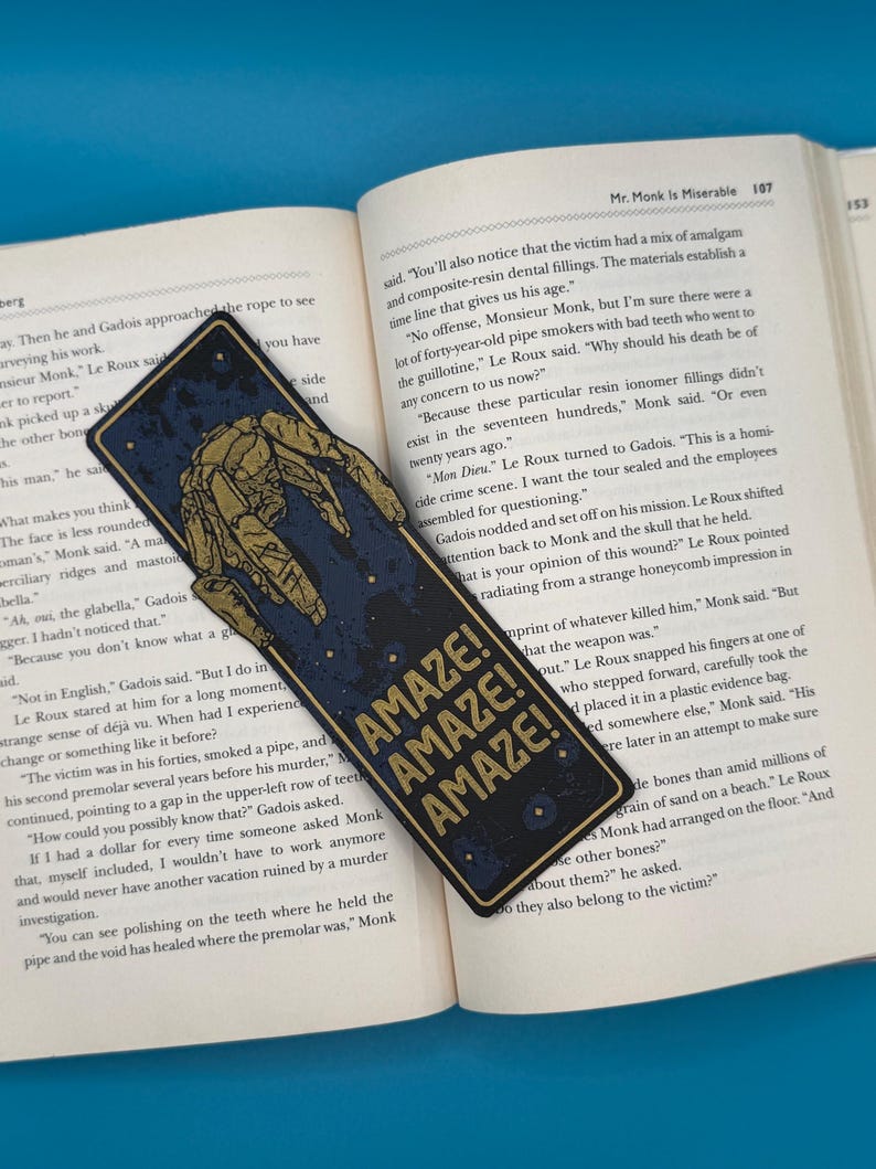 Rocky alien bookmark in dark blue with a gold mechanical outline and AMAZE AMAZE AMAZE text on an open sci fi book.