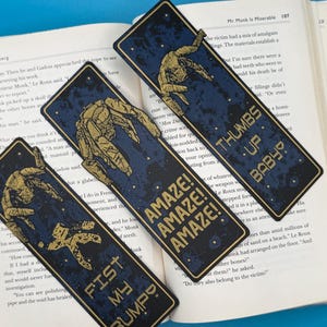 Three blue Rocky Alien bookmarks with gold mechanical art and Project Hail Mary-inspired sci-fi quotes: AMAZE! AMAZE! AMAZE!, THUMBS UP BABY, FIST MY BUMP.