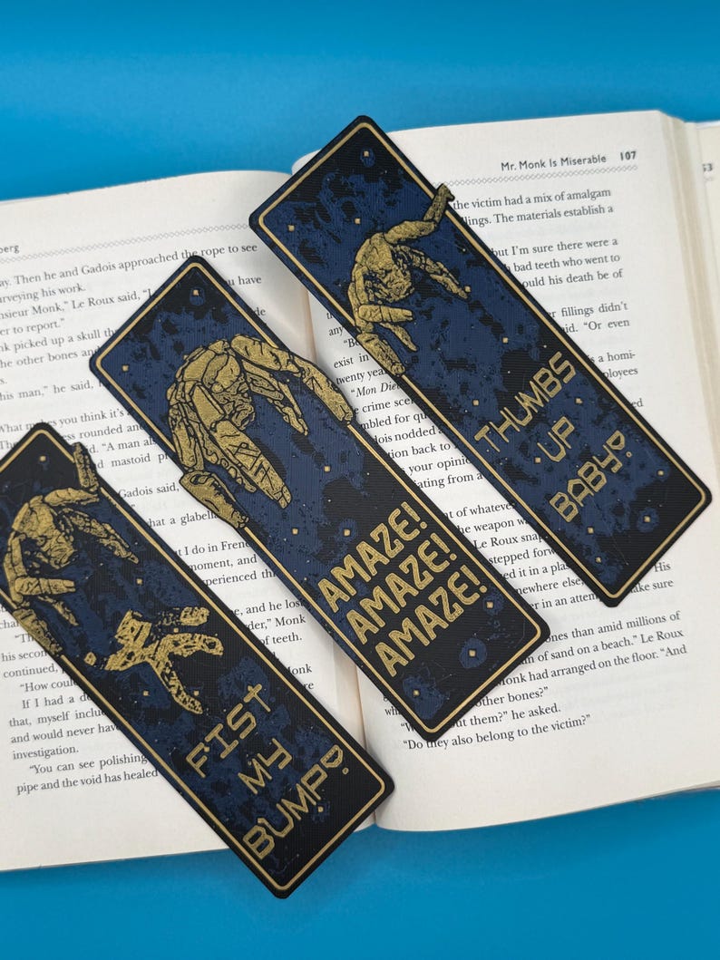 Three blue Rocky Alien bookmarks with gold mechanical art and Project Hail Mary-inspired sci-fi quotes: AMAZE! AMAZE! AMAZE!, THUMBS UP BABY, FIST MY BUMP.