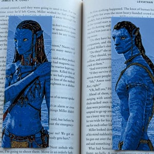 Könnte beinhalten: Ein Lesezeichen mit einem blauhäutigen Charakter aus dem Film Avatar. Der Charakter hat gelbe Augen und lange schwarze Haare. Das Lesezeichen befindet sich in einem Buch mit Text.