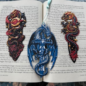 Puede incluir: Tres marcapáginas con forma de dragón en tonos rojos, negros y azules descansan sobre un libro abierto. Los marcapáginas presentan diseños intrincados, incluyendo alas y acentos de estrellas. Los marcapáginas están hechos de un material resistente y son ideales para los amantes de los libros de fantasía.