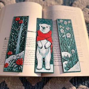 May include: Three decorative bookmarks with nature-themed designs. One features a polar bear wearing a red scarf, and the others depict floral and tree illustrations. The bookmarks are teal with white and red accents, resting on an open book.