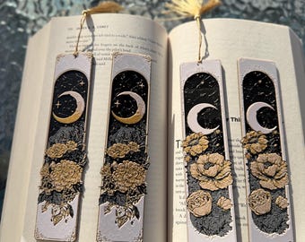 May include: Four rectangular bookmarks with a celestial design. Each bookmark features a crescent moon, stars, and gold-coloured floral accents against a black and white background. The bookmarks are displayed open in a book.