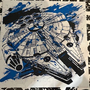 以下が含まれることがあります： ミレニアム・ファルコン宇宙船の様式化されたイラストが描かれた、完成したジグソーパズル。デザインは、白い背景に青と黒のアクセントを使用し、スプラッシュアート効果があります。パズルは正方形です。