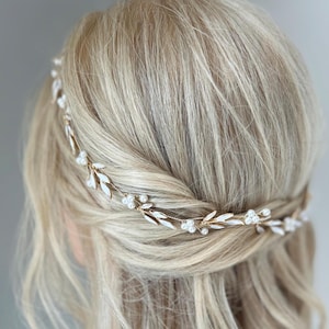 Puede incluir: Una guirnalda para el cabello dorada con detalles de hojas blancas y perlas. La delicada pieza para la cabeza se lleva en el cabello, con la parte posterior de la cabeza y el cabello rubio visibles. Es un accesorio decorativo.