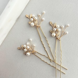 May include: Three gold hair pins with pearl and leaf embellishments. The pins are arranged in a criss-cross pattern.
