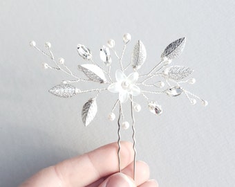 Puede incluir: Peine de pelo plateado con una flor blanca y hojas. El peine está decorado con pequeñas perlas y pedrería.