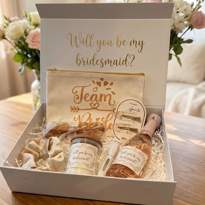 May include: A white gift box with a grey lid, filled with bridesmaid proposal gifts. The box contains a candle, a small bottle of champagne, a cosmetic bag, and hair accessories. The box lid reads "Will you be my bridesmaid?"