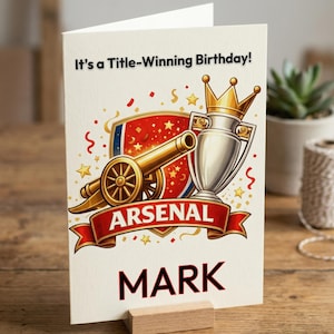 May include: A birthday card with an Arsenal theme. The card features a cannon, trophy, and the text "It's a Title-Winning Birthday!" with the name "MARK" below. The card is propped up on a wooden stand.