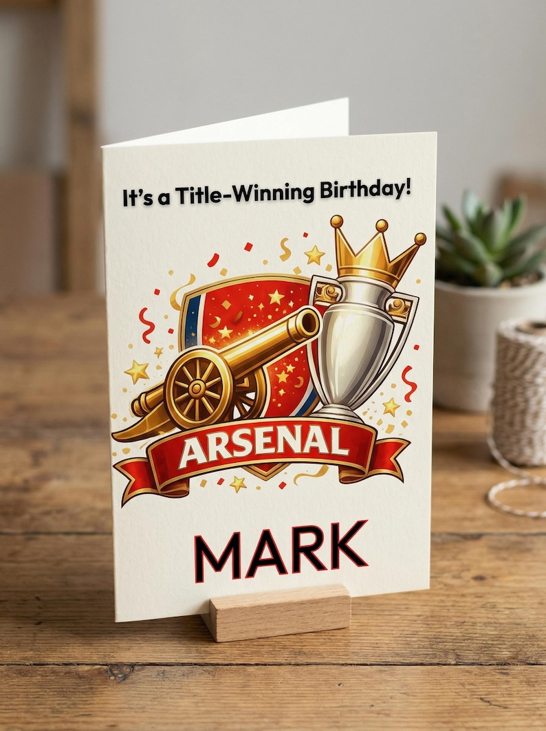 May include: A birthday card with an Arsenal theme. The card features a cannon, trophy, and the text "It's a Title-Winning Birthday!" with the name "MARK" below. The card is propped up on a wooden stand.