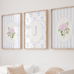 May include: Three framed wall art prints featuring light blue vertical stripes and oval centres. The outer prints display pink hydrangea illustrations. The centre print features a blue letter J surrounded by a blue lattice pattern with small blue flowers.