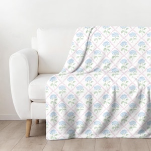 May include: A white throw blanket draped over a white sofa. The blanket features a repeating pattern of blue hydrangea flowers and green leaves within a pink lattice design. The sofa has wooden legs.