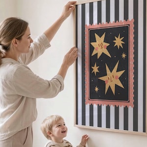 May include: A rectangular wall art piece in a light wood frame featuring a black and white vertical striped background. The centre displays a black rectangle with a pink scalloped border containing yellow star illustrations with smiling faces.