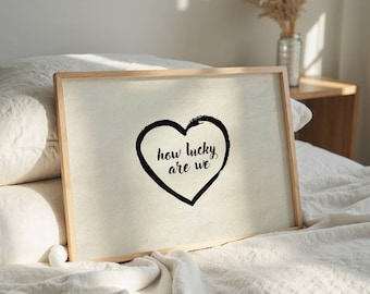 May include: A framed print with a light beige background and a black heart design. The heart contains the handwritten text "how lucky are we". The frame is made of light-colored wood. The print is displayed on a bed with white bedding.