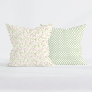 May include: Two square throw pillows. The left pillow features a repeating pattern of light pink and green abstract botanical shapes on a white background. The right pillow displays a light green and white gingham check pattern throughout.