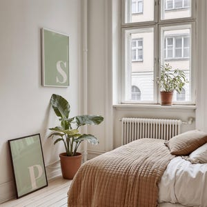 May include: Two rectangular wall art prints featuring a sage green background and a large white letter. One print displays the letter S and is mounted on the wall, while the other print displays the letter P and rests on the floor.