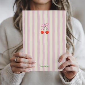 Puede incluir: Un cuaderno con un diseño de rayas crema y rosa. La portada presenta un par de cerezas rojas con un lazo rosa. El nombre "Victoria Hart" está impreso en la parte inferior de la portada.