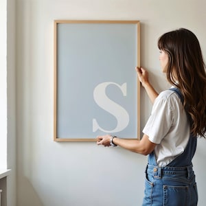 May include: A rectangular wall art print featuring a large, white capital letter S centered on a solid light blue background, enclosed within a thin, light-toned wooden frame. The design is clean and simple, focusing on the typography.