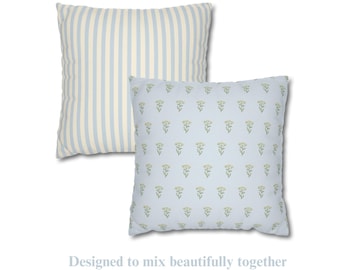 May include: Two square throw pillows. One pillow features a light blue and cream vertical striped pattern. The second pillow displays a light blue background with a repeating small green floral print. The text below reads: Designed to mix beautifully together. Cozy, timeless, and easy to layer.