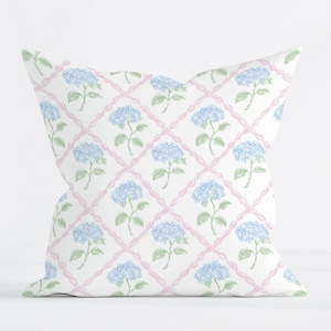 May include: Square throw pillow with a white background featuring a repeating pattern of blue hydrangea flowers and green leaves. The flowers are set within a pink lattice design. The pillow is a decorative home accent.