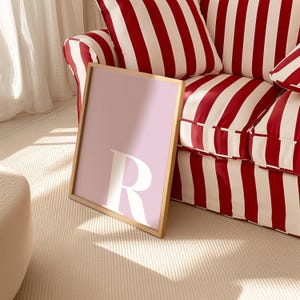 May include: A rectangular framed wall art piece featuring a light lavender background with a large, bold, white capital letter R centered in the middle. The frame is a thin, light-toned wood, and the artwork is displayed in a vertical orientation.