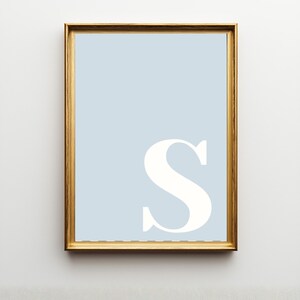 Pode incluir: Uma impress&atilde;o de arte de parede retangular vertical com uma letra S mai&uacute;scula grande, branca e em estilo serifado, centralizada num fundo azul-claro s&oacute;lido. A impress&atilde;o est&aacute; emoldurada por uma moldura fina de metal dourado polido.