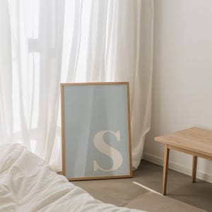 May include: A rectangular wall art print in a light wooden frame featuring a large, off-white capital letter S centred on a muted, pale blue background. The artwork is displayed in a vertical orientation with a clean, simple design aesthetic.