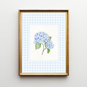 May include: A framed watercolor print featuring a cluster of light blue hydrangeas with green leaves. The artwork is set against a light blue and white gingham pattern, enclosed in a gold frame.