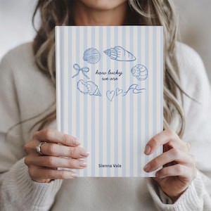 May include: A hardcover notebook with a blue and white striped cover. The cover features blue seashell illustrations and the text "how lucky we are" and "Sienna Vale".