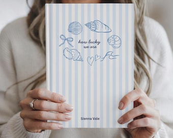 Könnte beinhalten: Ein Notizbuch mit blau-weiß gestreiftem Einband. Der Einband zeigt blaue Muscheln und den Text "how lucky we are" und "Sienna Vale".