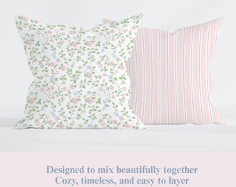 May include: Two decorative throw pillows. One pillow features a white background with a floral pattern of pink and blue flowers and green leaves. The other pillow has pink and green vertical stripes. The text at the bottom says, "Designed to mix beautifully together. Cozy, timeless, and easy to layer."