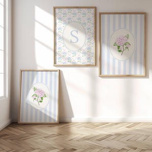 Op de afbeelding: Drie rechthoekige wandkunstprints in licht houten lijsten. Eén print heeft een blauw en roze ruitpatroon met een centrale ovaal met de letter S. Twee prints hebben lichtblauwe verticale strepen met een centrale ovaal met roze hortensia's.