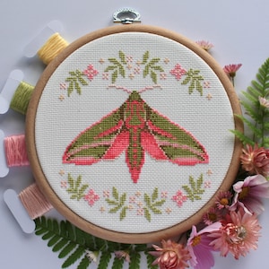 May include: A completed cross-stitch embroidery of a moth with pink and green wings, set in a wooden hoop. The moth is surrounded by a floral design. Spools of thread and flowers are arranged around the hoop.